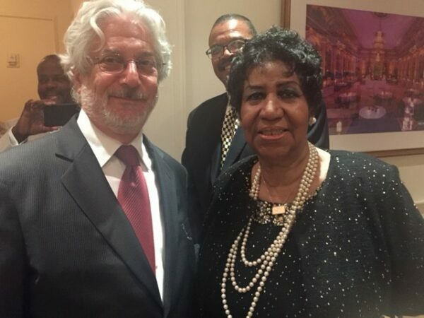 A white haired man with a suit stands next to a black haired woman in a dress